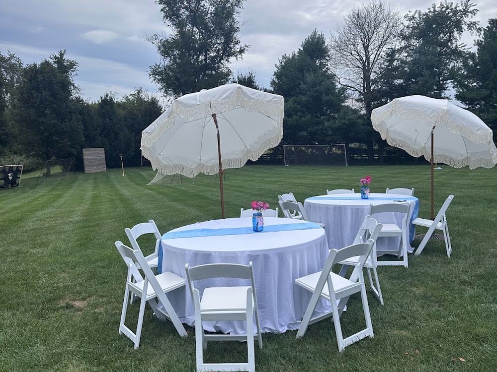 A round table with a floor length linen, white folding chairs and a fringed umbrella on a lawn