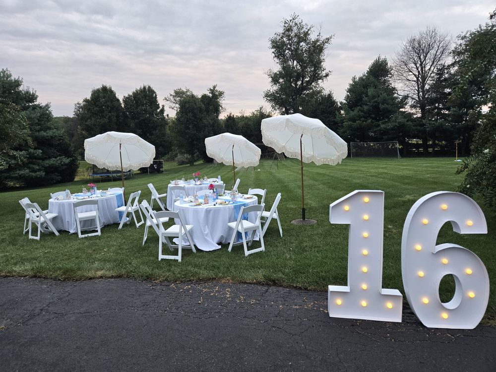 Round tables, white chairs and three umbrellas on a lawn at dusk with a lit marquee number 16